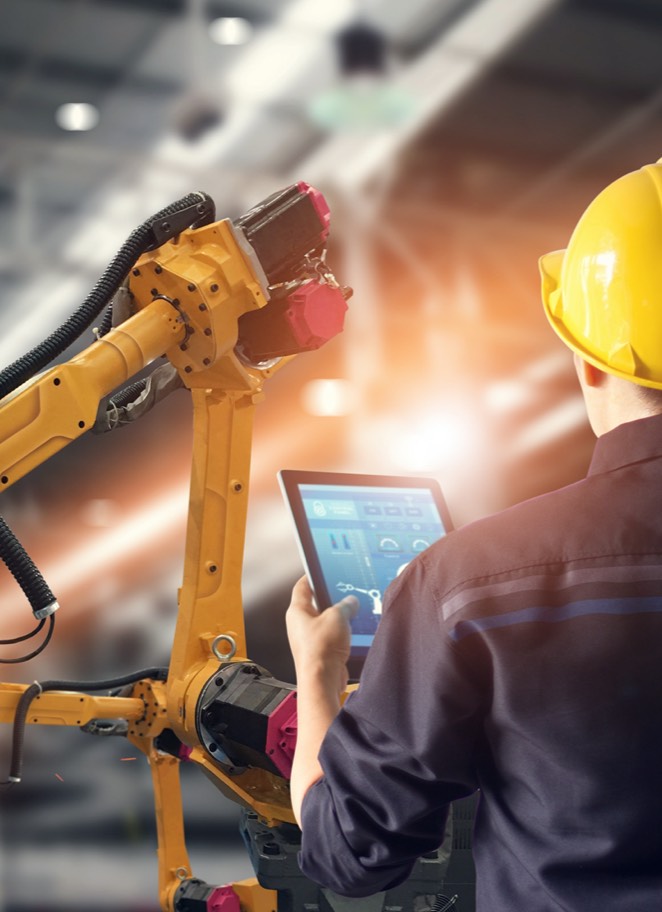 A view from behind of someone wearing a hardhat and holding a digital tablet standing in front of a piece of machinery